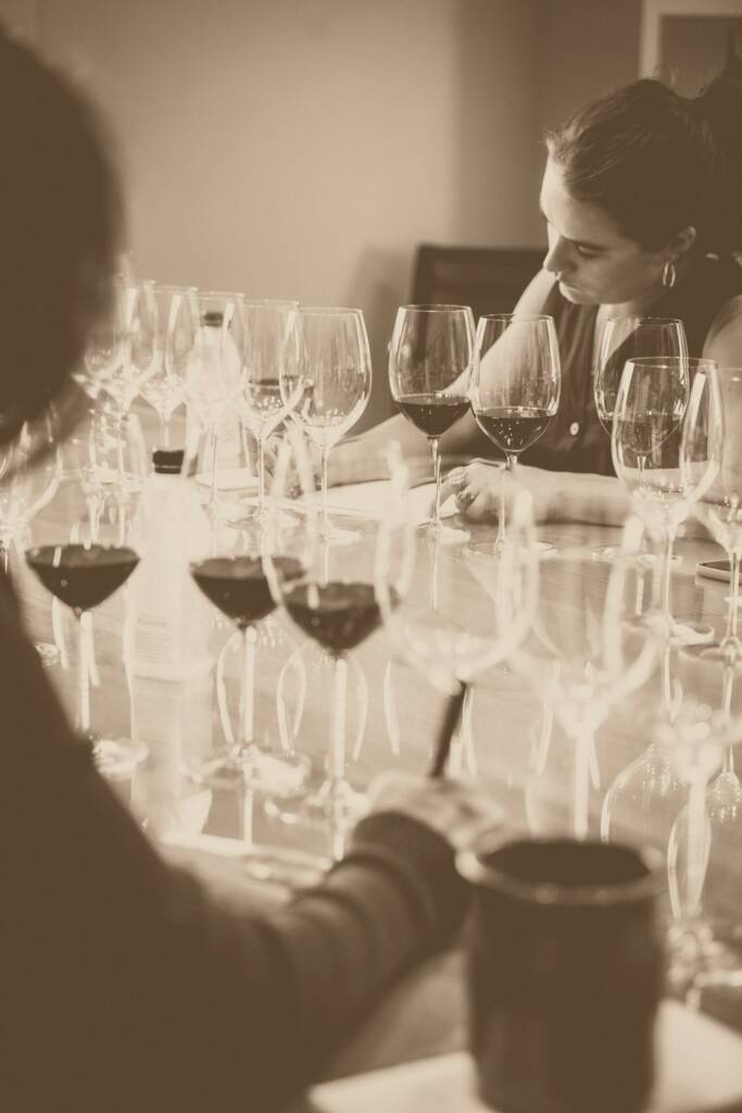 Alexandra Evnin and Michel Rolland sit at a table with various wine glasses, engaged in tasting and taking notes. The scene is captured in a sepia-toned photograph.
